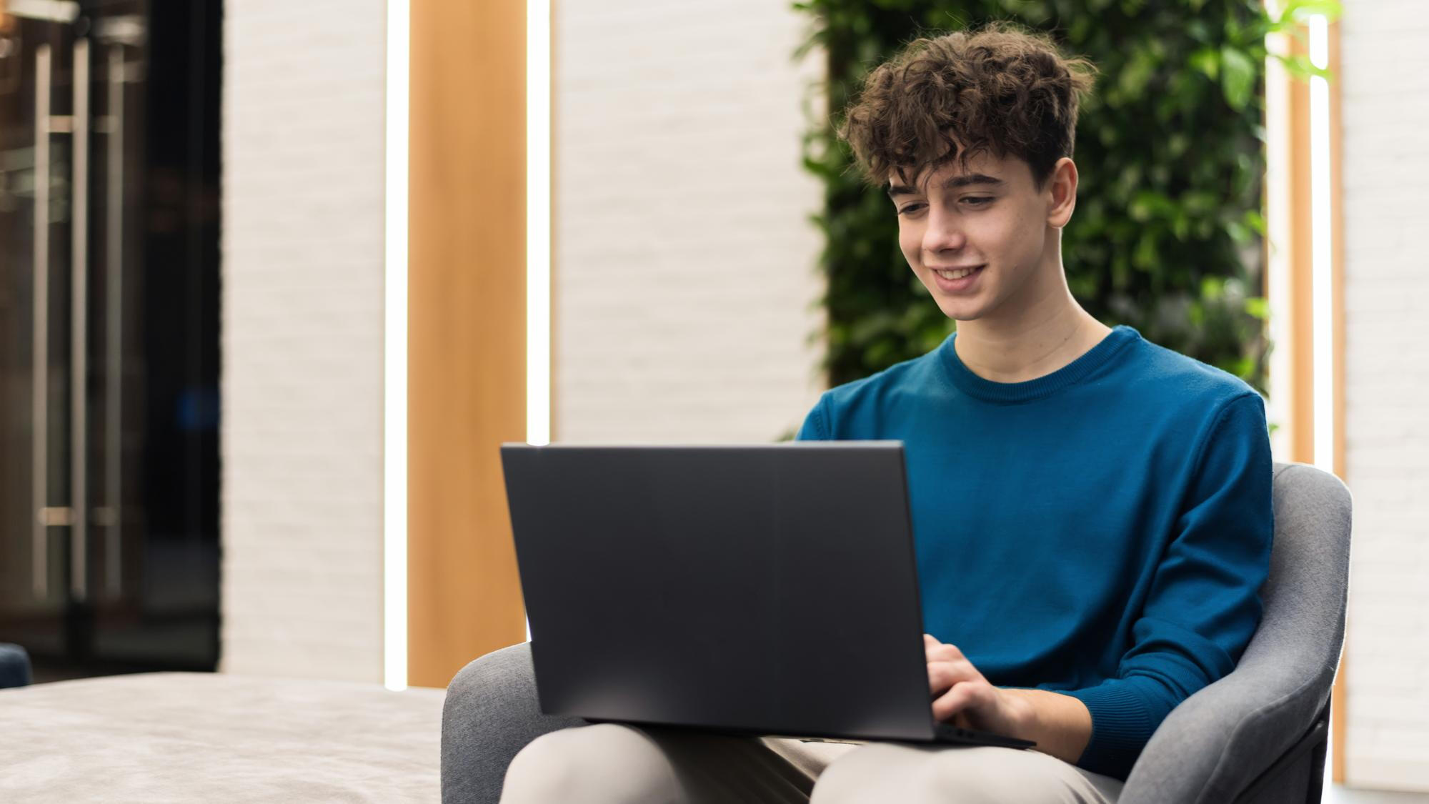 A young man in a chair, engaged with his laptop, displaying a relaxed and studious demeanor.