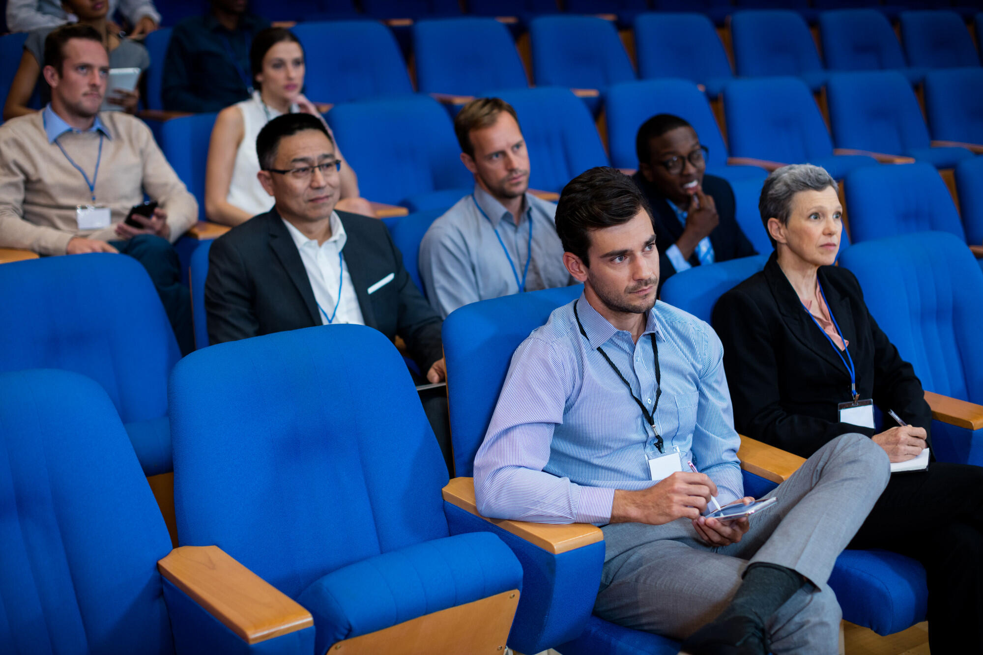 Audience members seated in a lecture hall, wearing badges and taking notes during a conference or seminar.