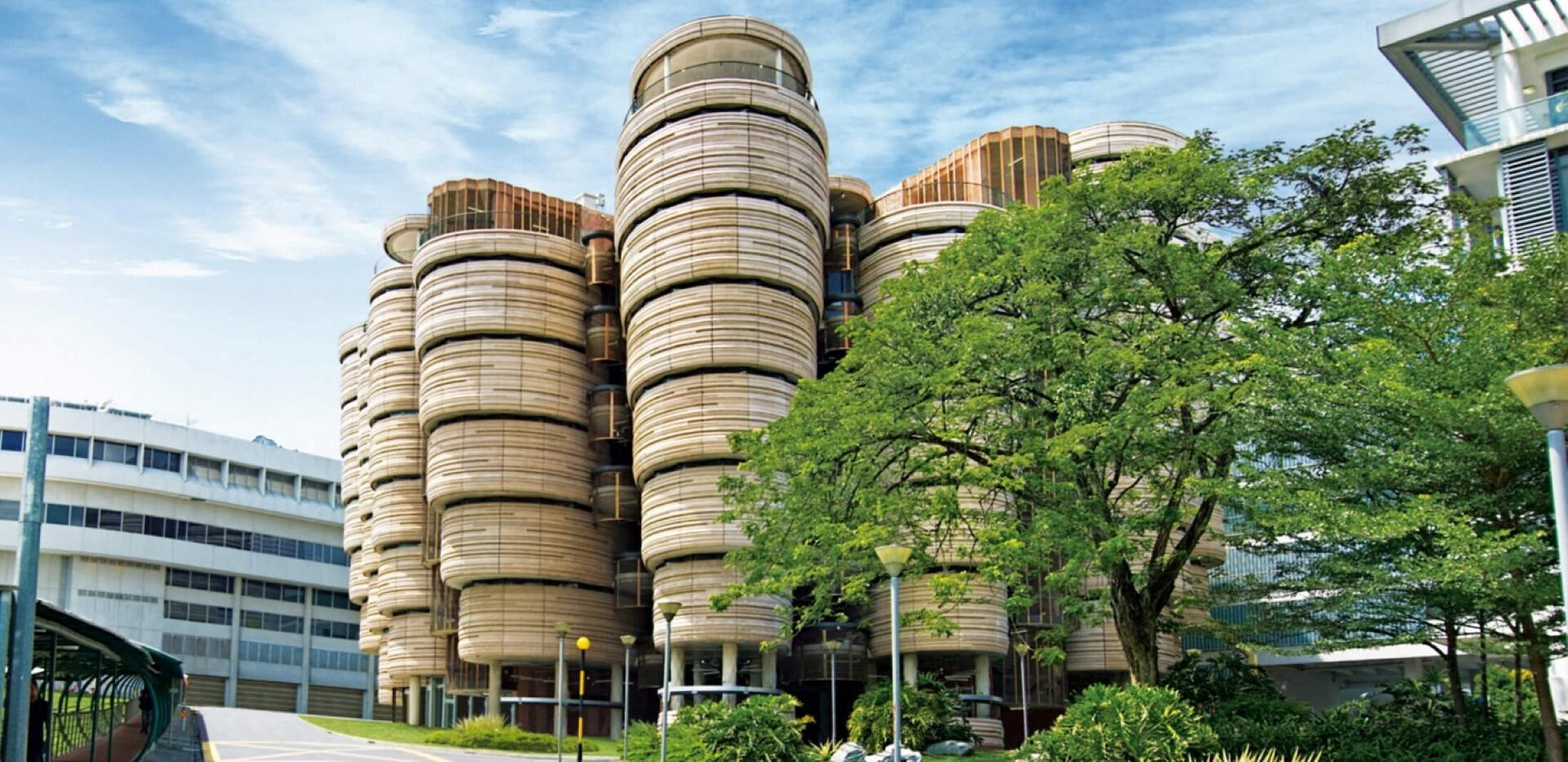 Modern cylindrical building with layered, rounded wooden panels, surrounded by trees and nearby urban structures under a partly cloudy sky.