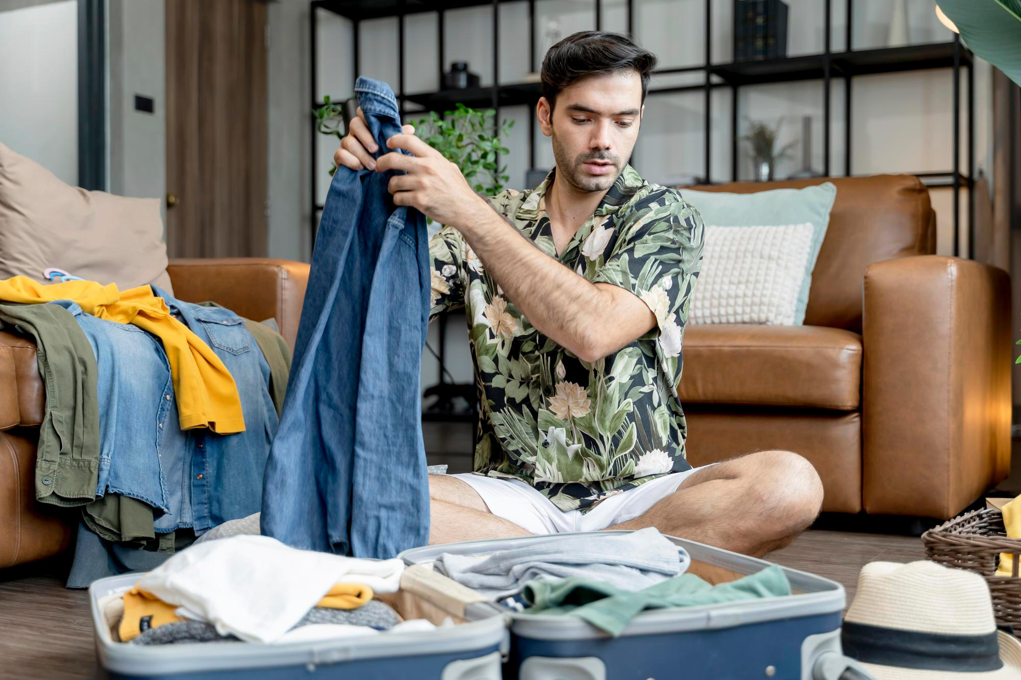 Man sitting on floor packing clothes into suitcase in living room with couch and pillows in background.