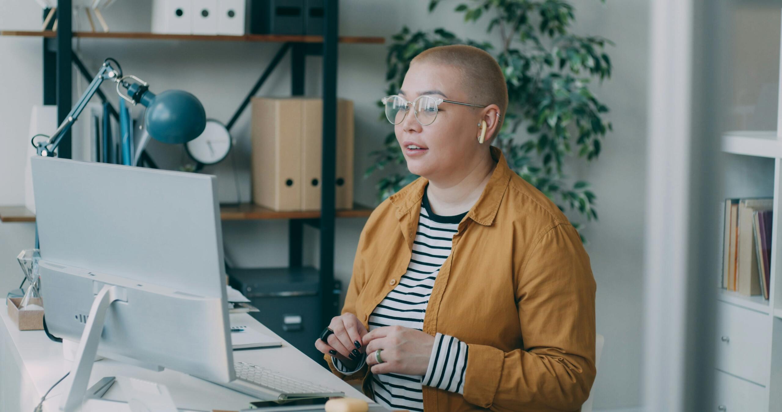 A woman wearing glasses sits at a desk, focused on her computer screen, with a notepad beside her.