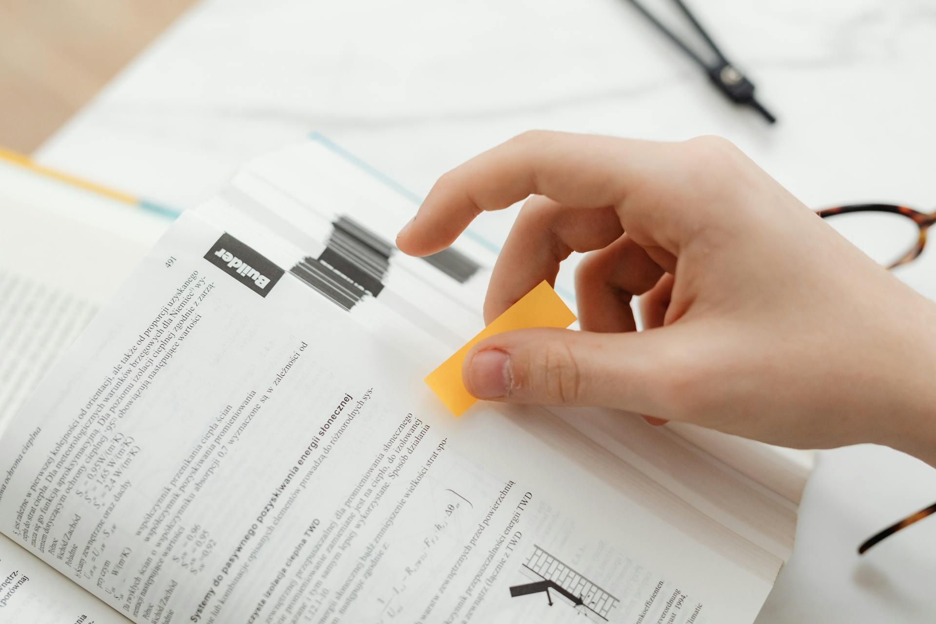 A hand places a sticky note in an open book, symbolizing studying or learning