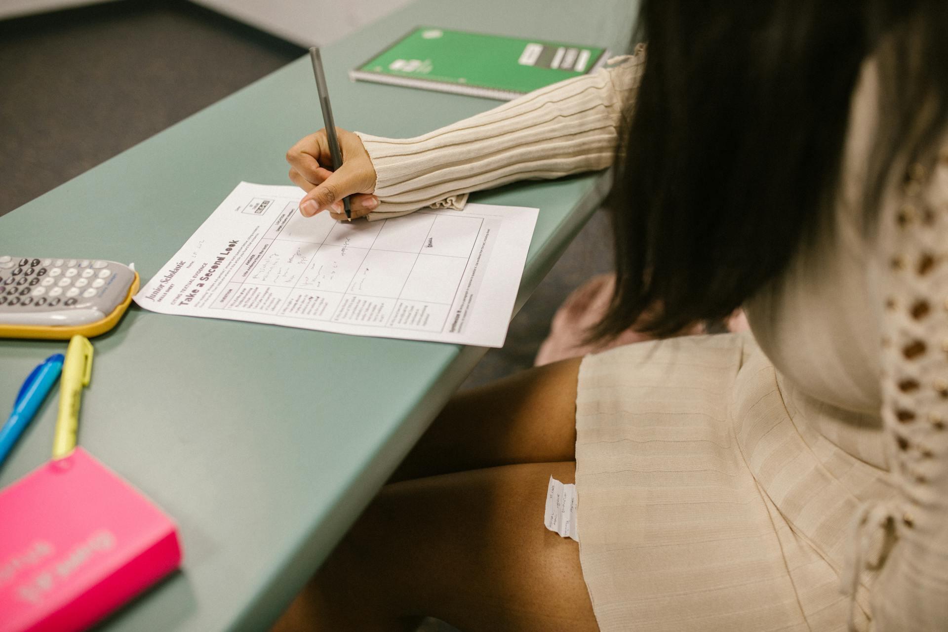 A person sitting at a desk, writing on a document with a pen, while various stationery items are visible, including a calculator and highlighters.
