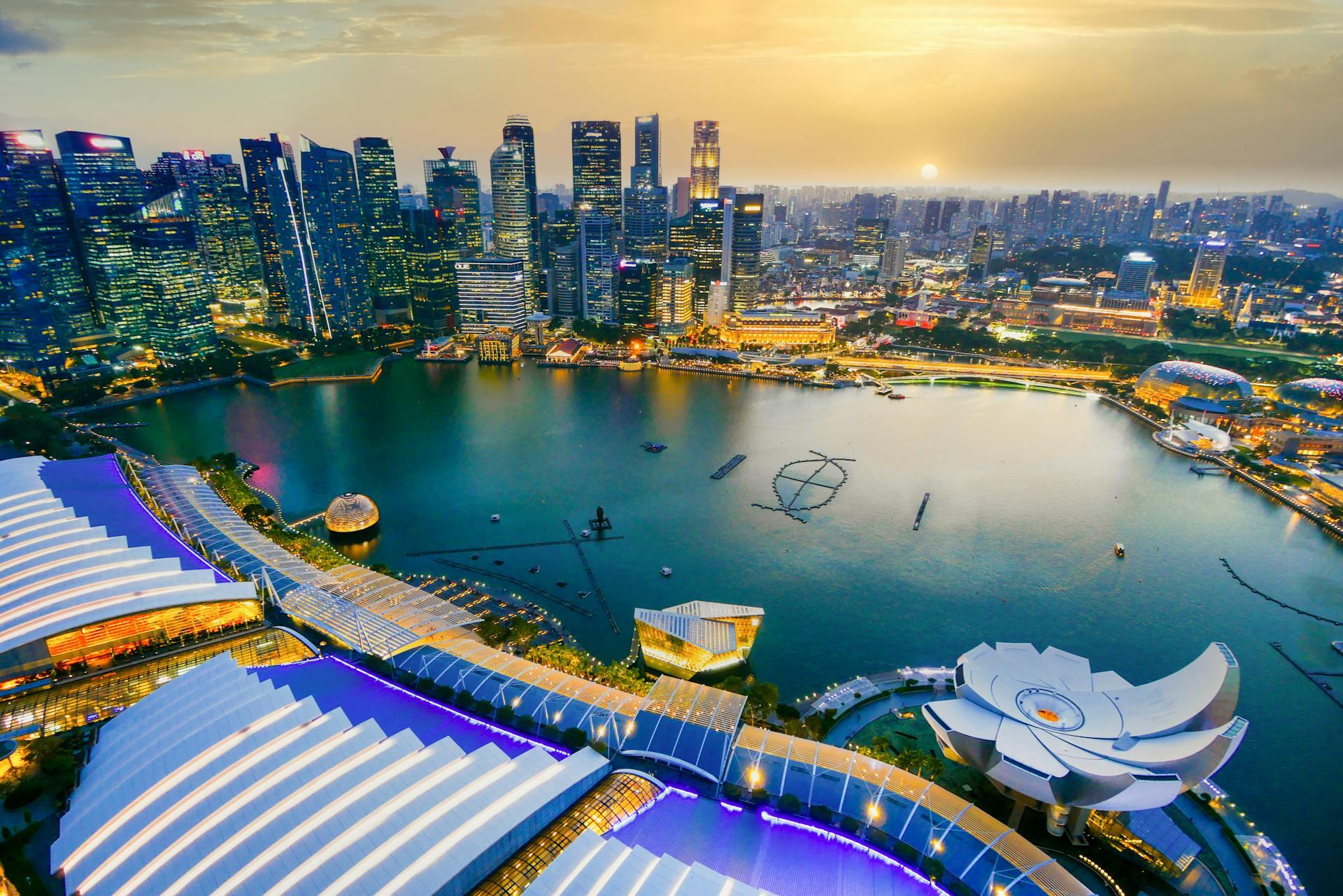A stunning aerial view of Singapore's Marina Bay at sunset. Skyscrapers illuminate the skyline, while futuristic buildings curve along the waterfront.