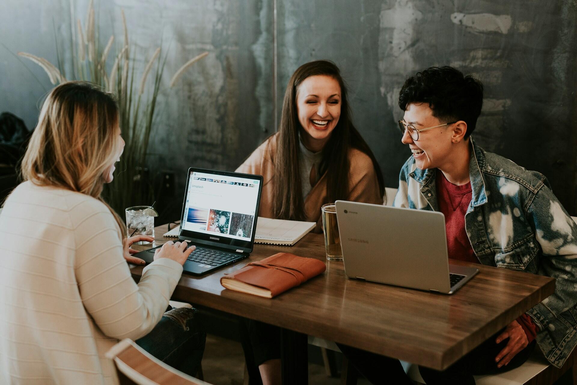 Three people laughing around a table with laptops and notebooks