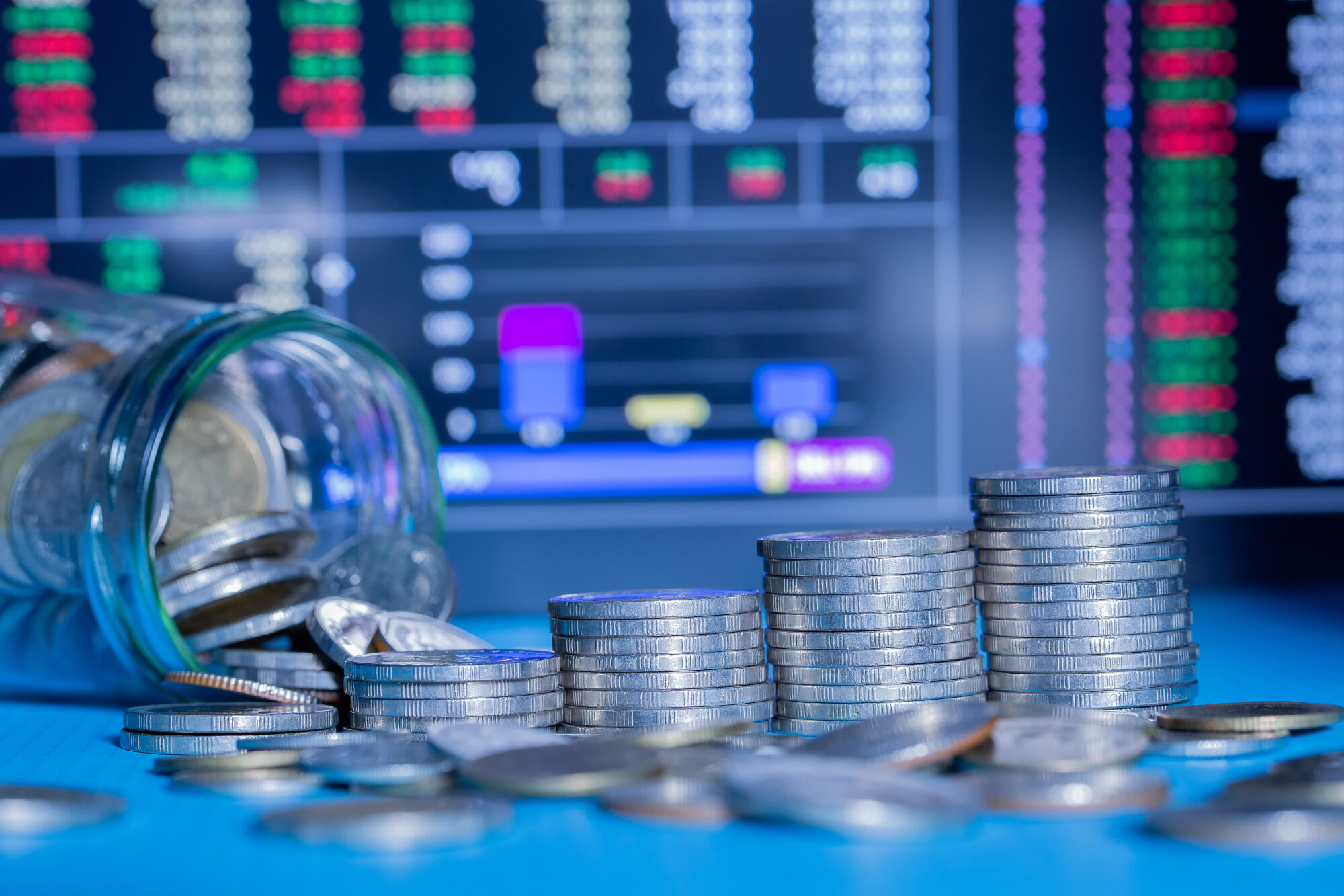 A jar filled with coins sits on a table next to a stock market chart, symbolizing investment and savings.