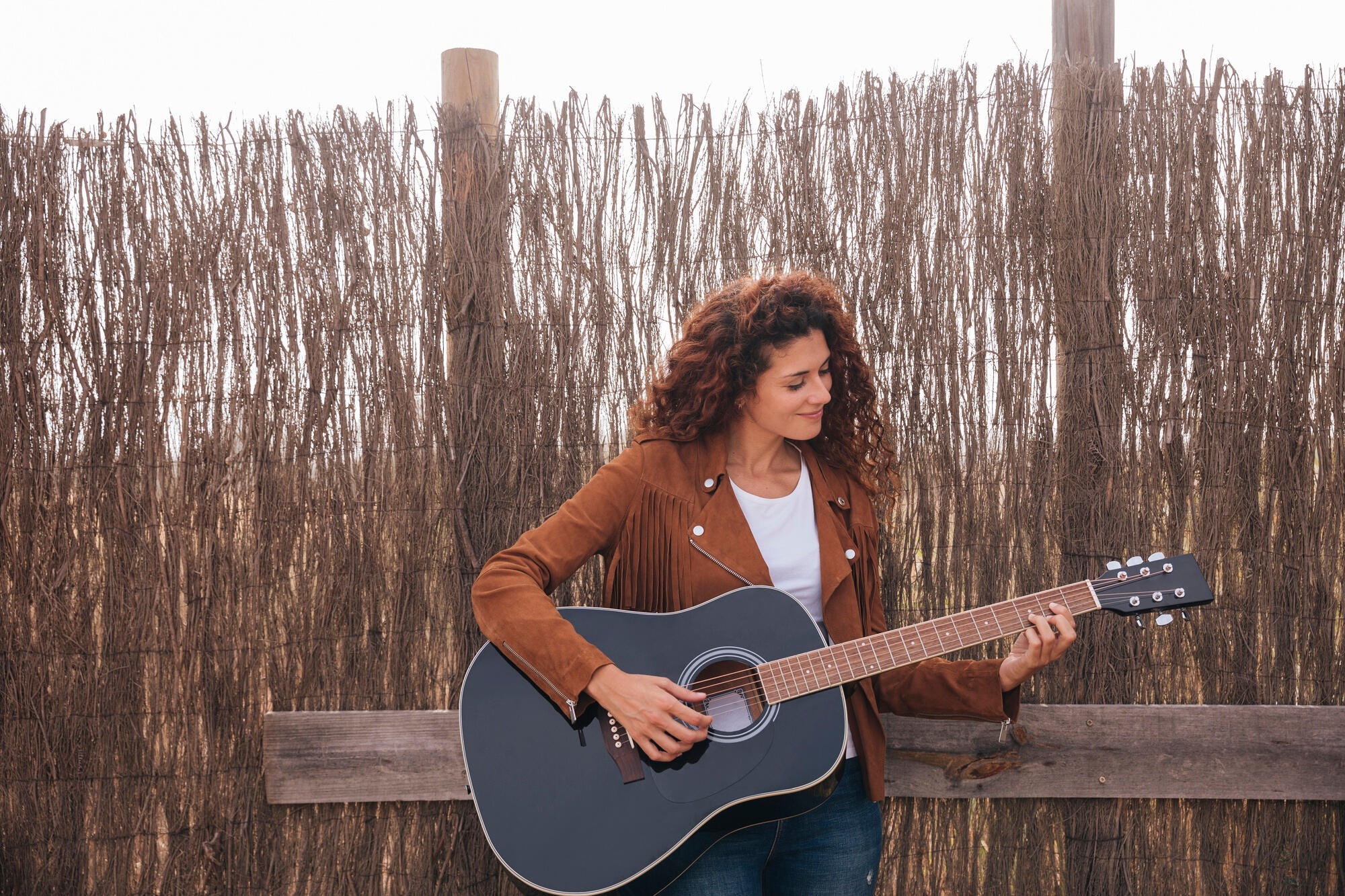 A woman with curly hair strums an acoustic guitar, focused on her music in a cozy setting.