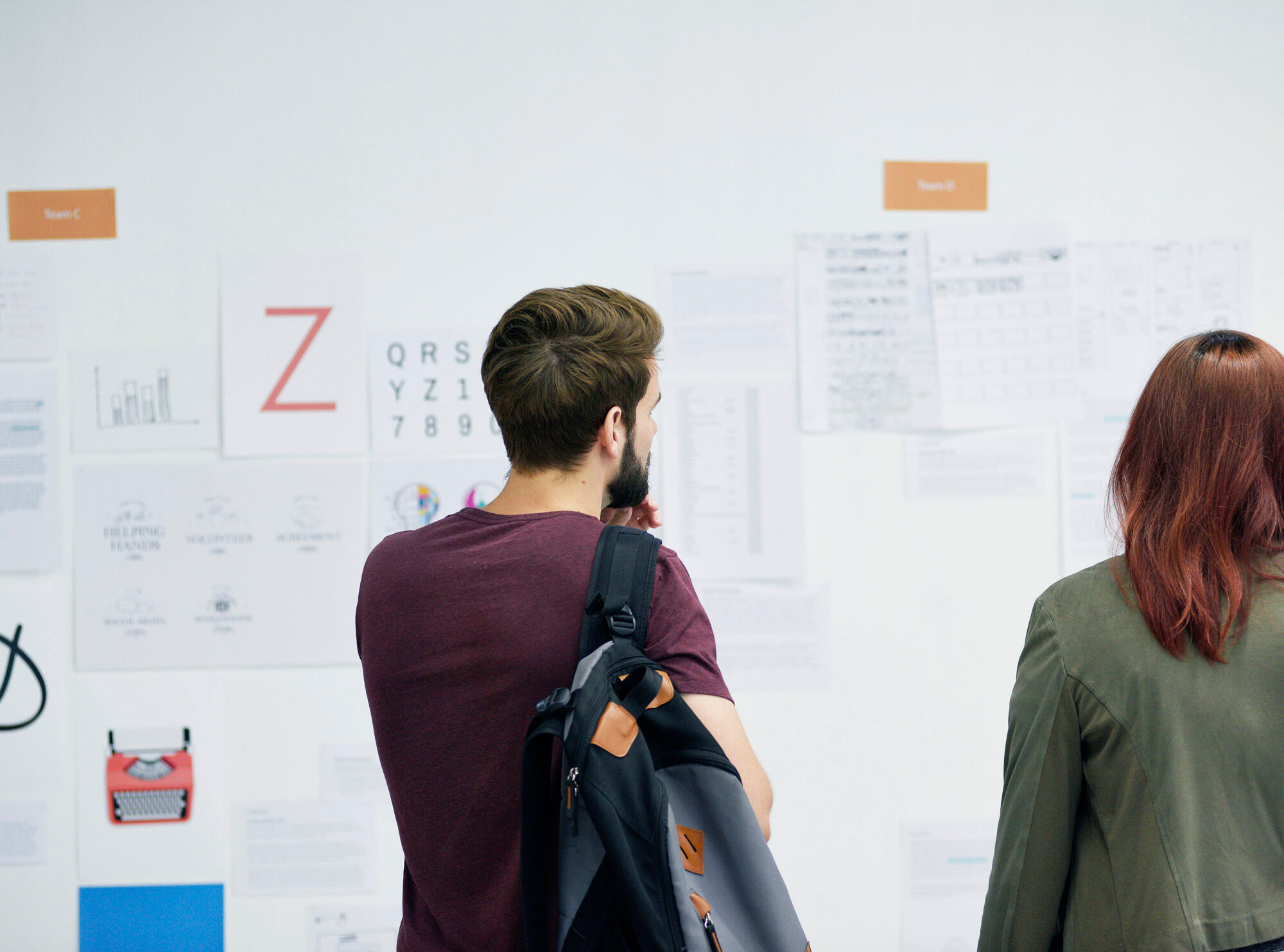 Two people with backpacks stand facing a wall covered with charts, graphs, and documents labeled Team C and Team D.