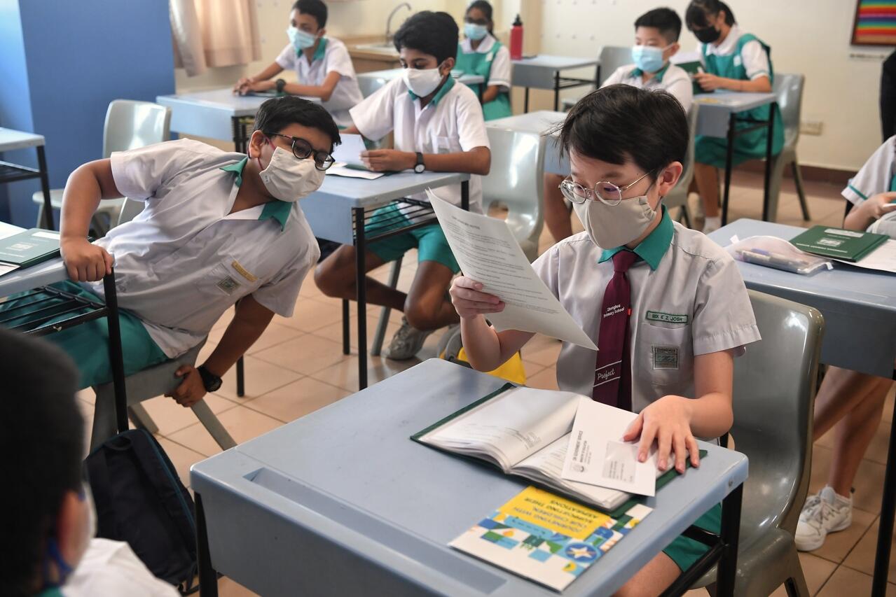 Students wearing masks sitting in a classroom, with one student reading a document and another student leaning back in their chair.