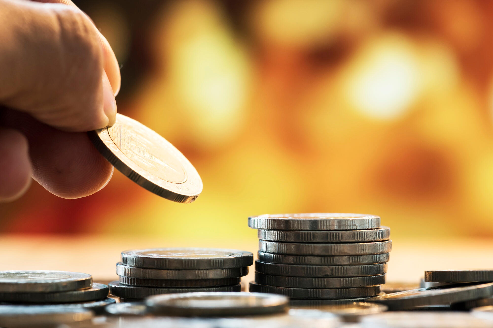 A person holds a single coin above a larger pile of assorted coins on a flat surface.
