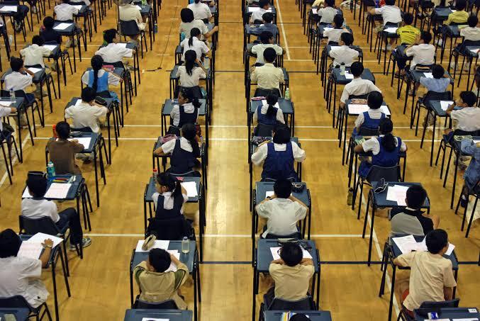 A large group of students sitting at desks in a classroom during an exam, with their heads down and papers in front of them.