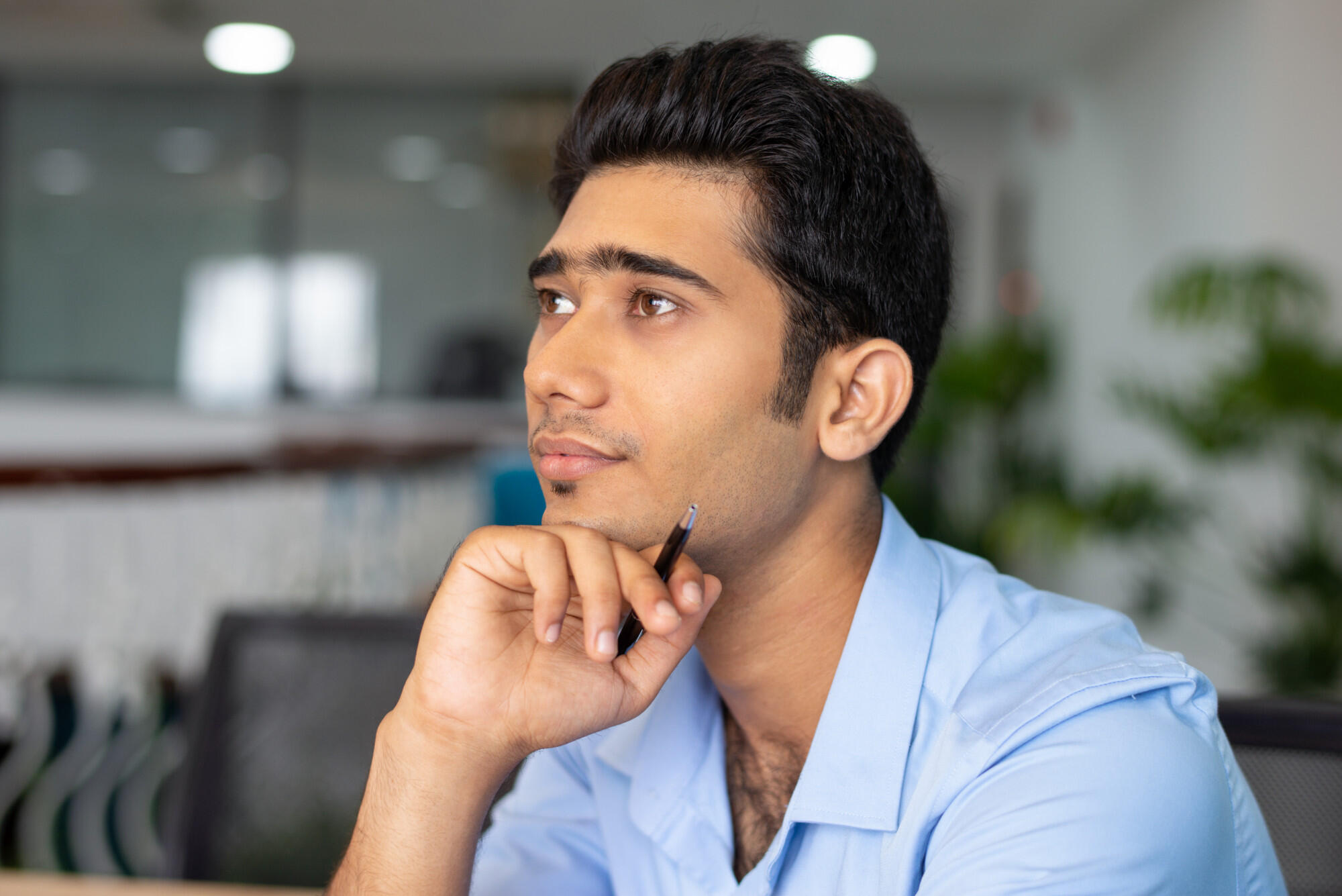 Young man in a blue shirt holding a pen, looking thoughtfully to the side in a modern office setting.