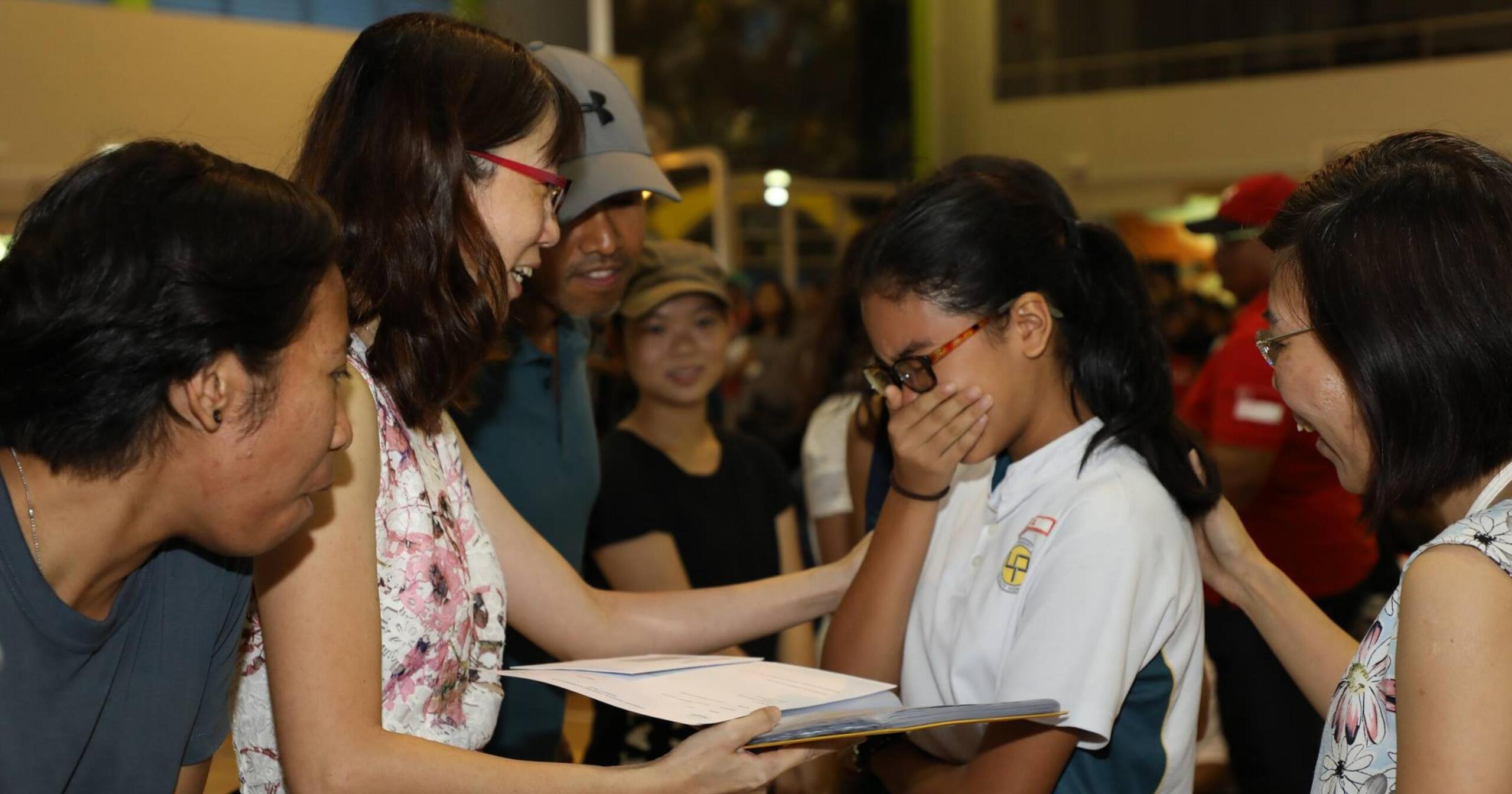 A group of people, including a girl in a school uniform, reacts emotionally while receiving her PSLE results in a crowded indoor setting.