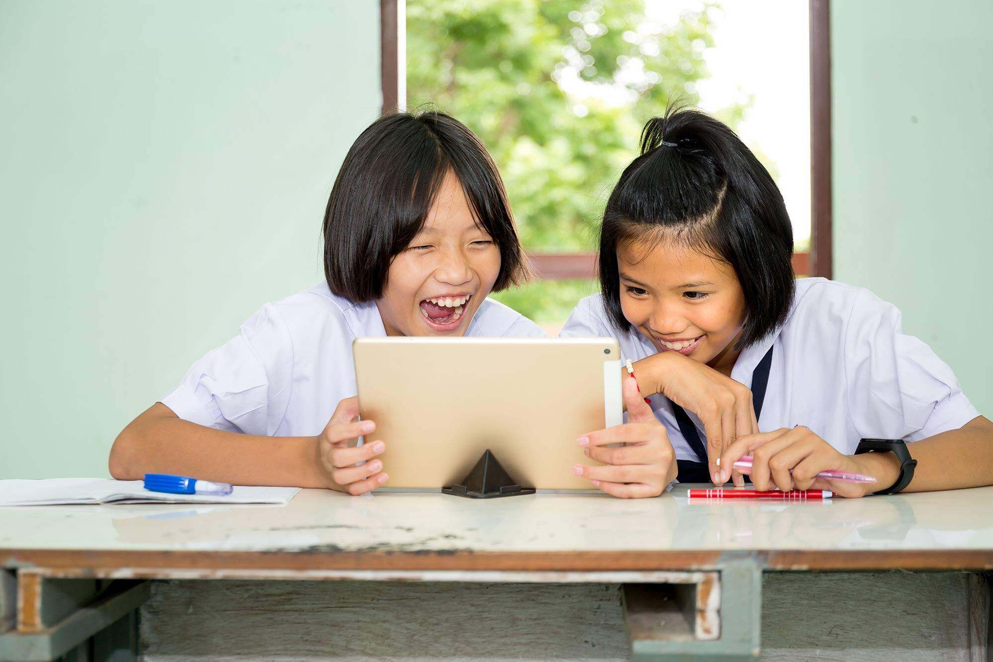 Two young girls, sitting at a table in a classroom, joyfully looking at a tablet together.
