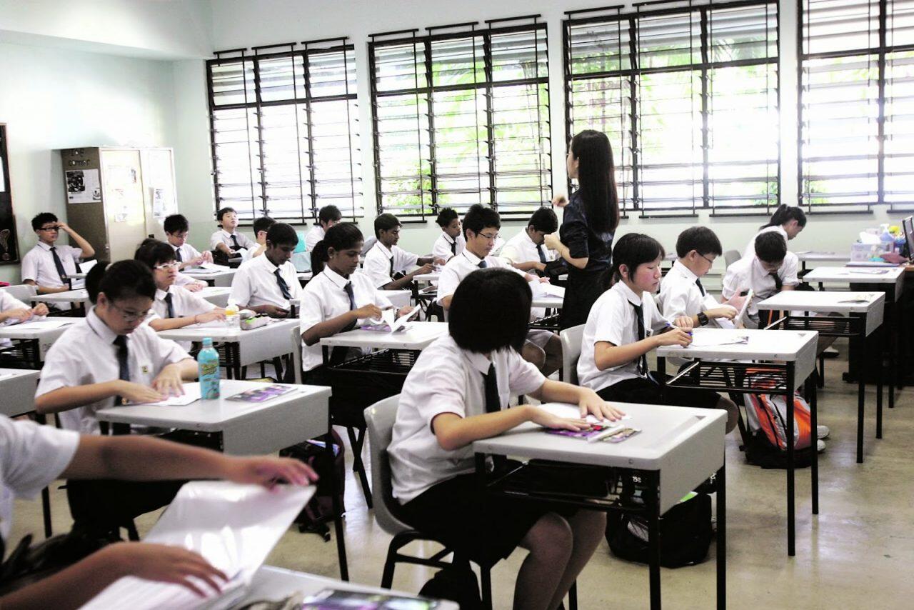 A classroom filled with students sitting at desks, engaged in various activities while a teacher stands at the front.