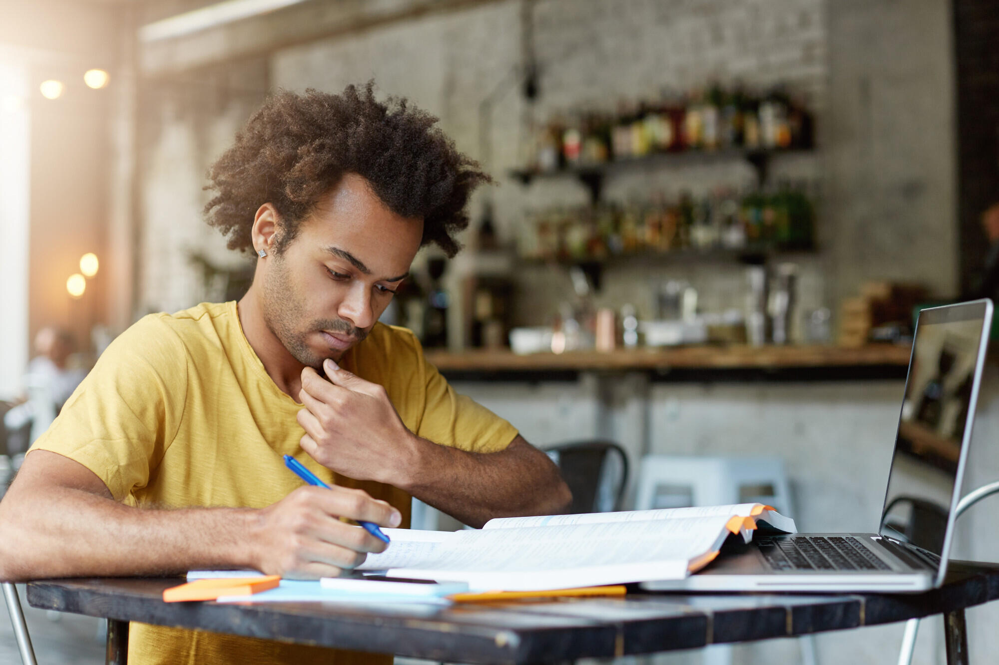 A man seated at a table, using a laptop and holding a pen, focused on his work.
