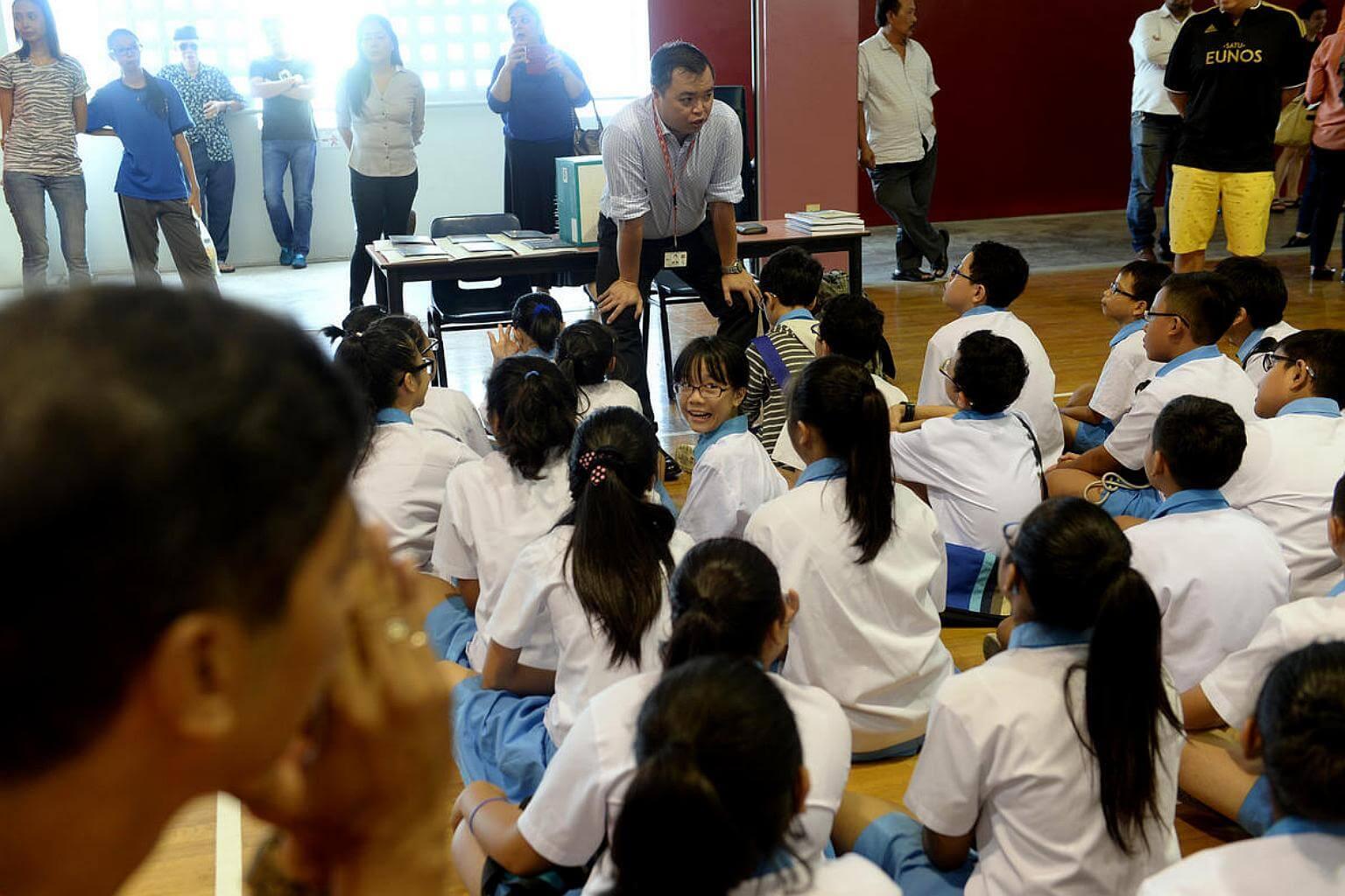 A group of children sitting on the floor, while a teacher stands and talks to them. Adults are in the background.