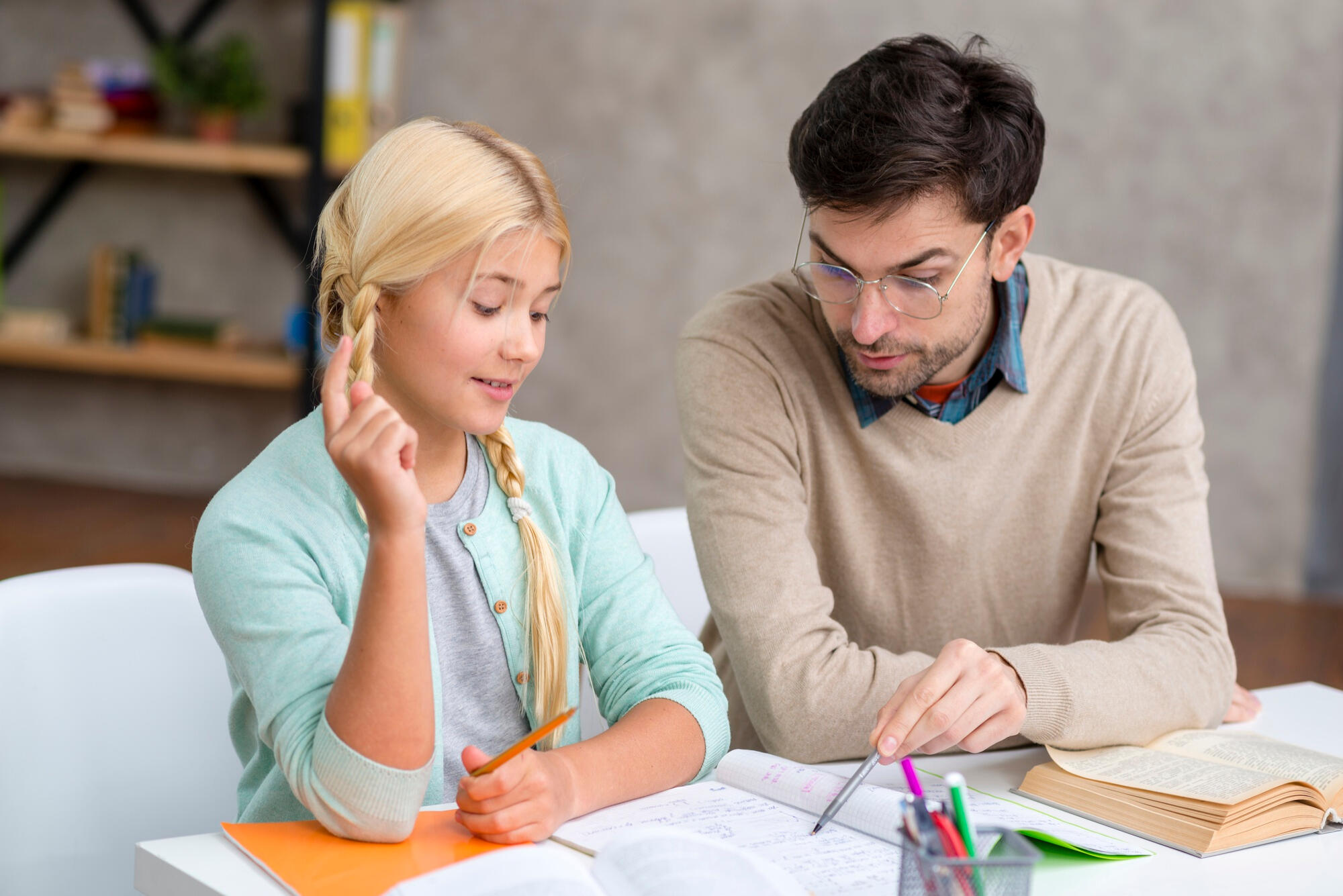 A man and a young girl sit at a table, surrounded by books, engaged in reading and discussion.