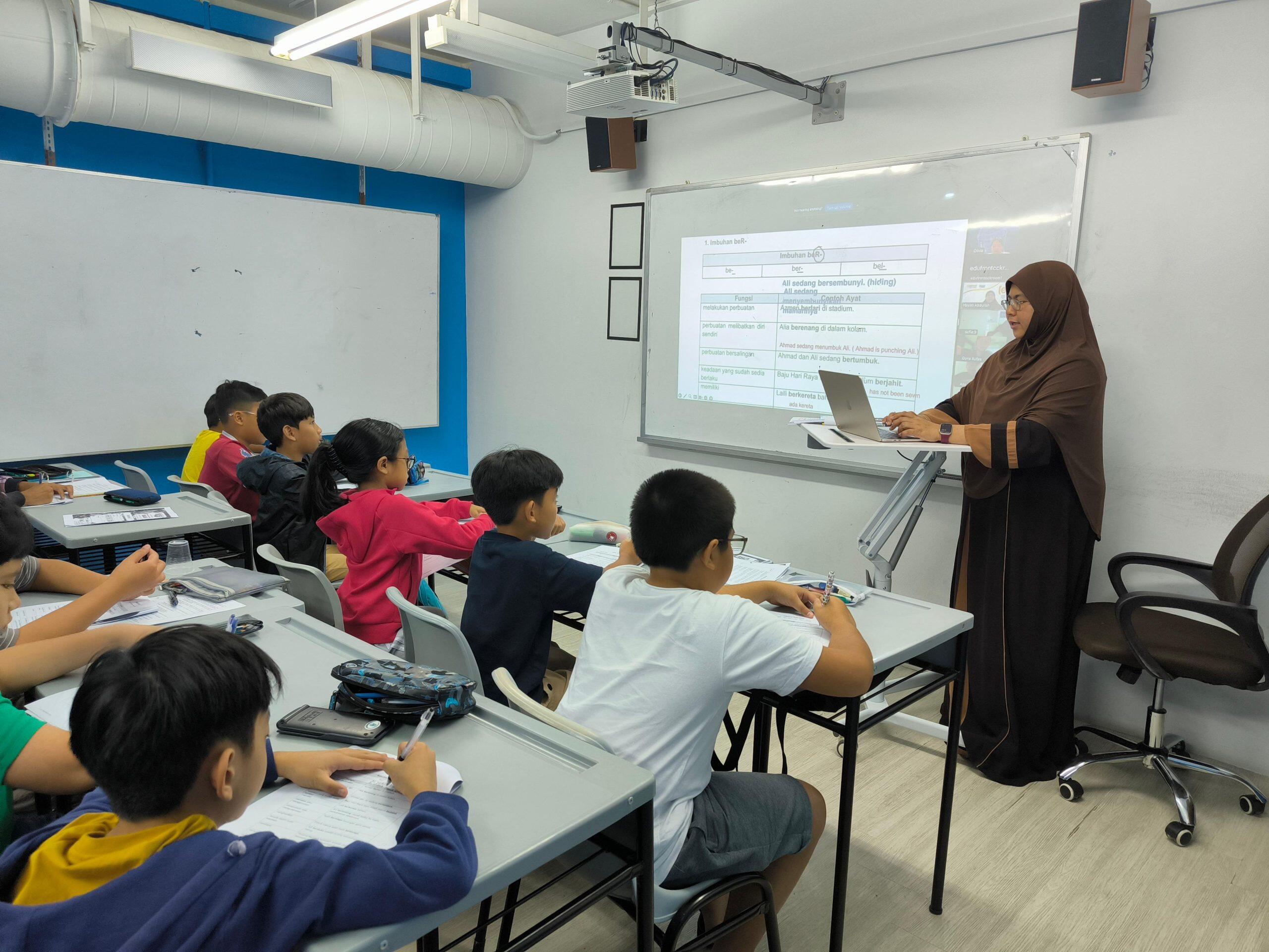 A teacher is teaching a class of students in a classroom setting. Students are sitting at desks facing the teacher, who is standing in front of a whiteboard with a laptop and teaching materials.
