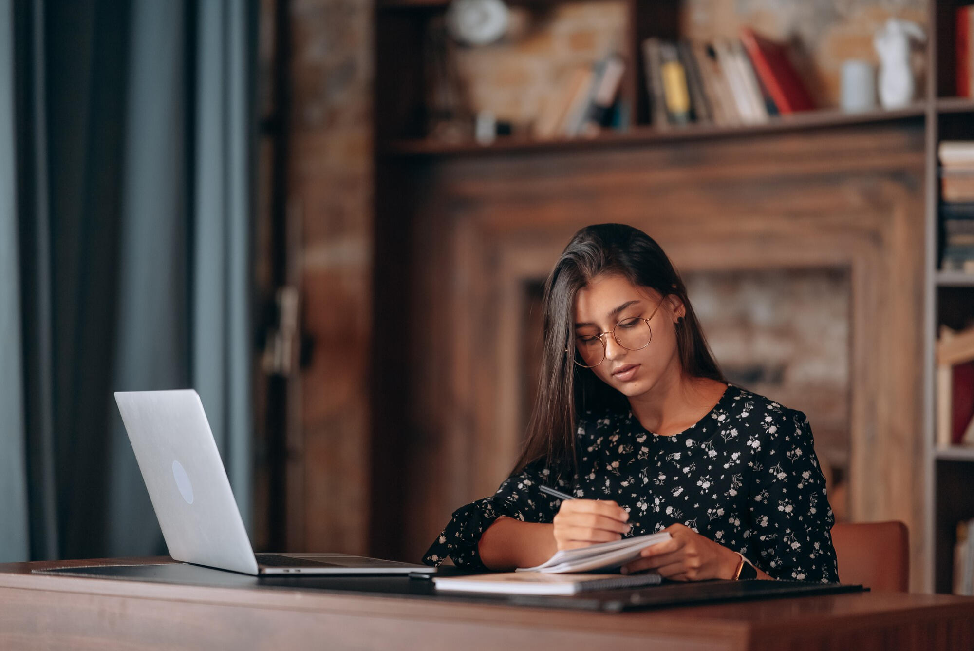 Young woman with glasses writing in a notebook at a wooden desk with a laptop in a cozy, book-filled room.