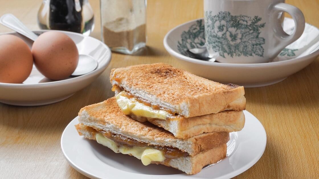 Golden toasted cheese sandwiches stacked on a white plate beside eggs and a floral teacup, creating a warm, rustic breakfast scene.