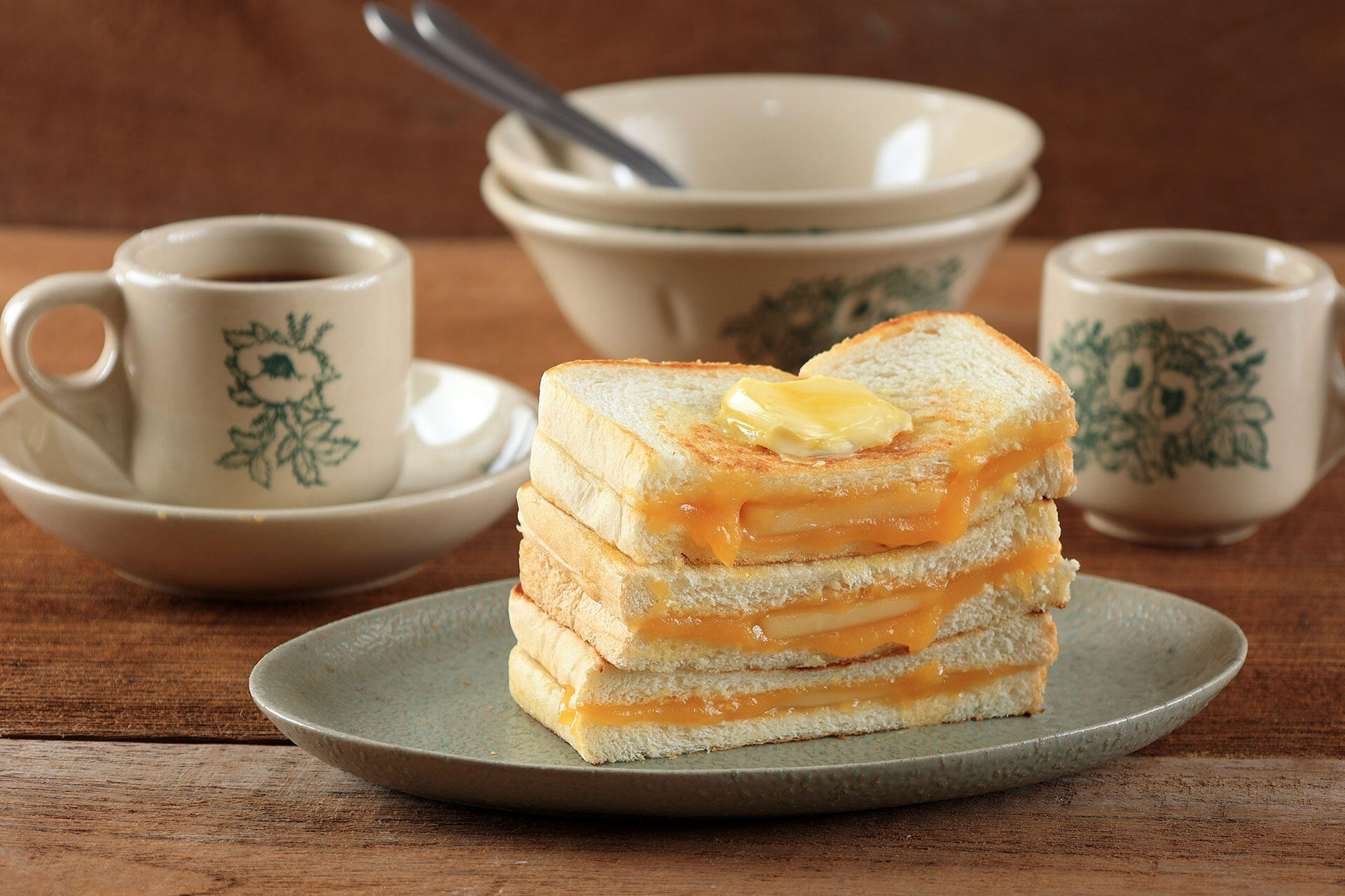 Stack of golden toasted cheese sandwiches on a ceramic plate, with butter melting on top, vintage cups and bowl in a warm rustic setting.