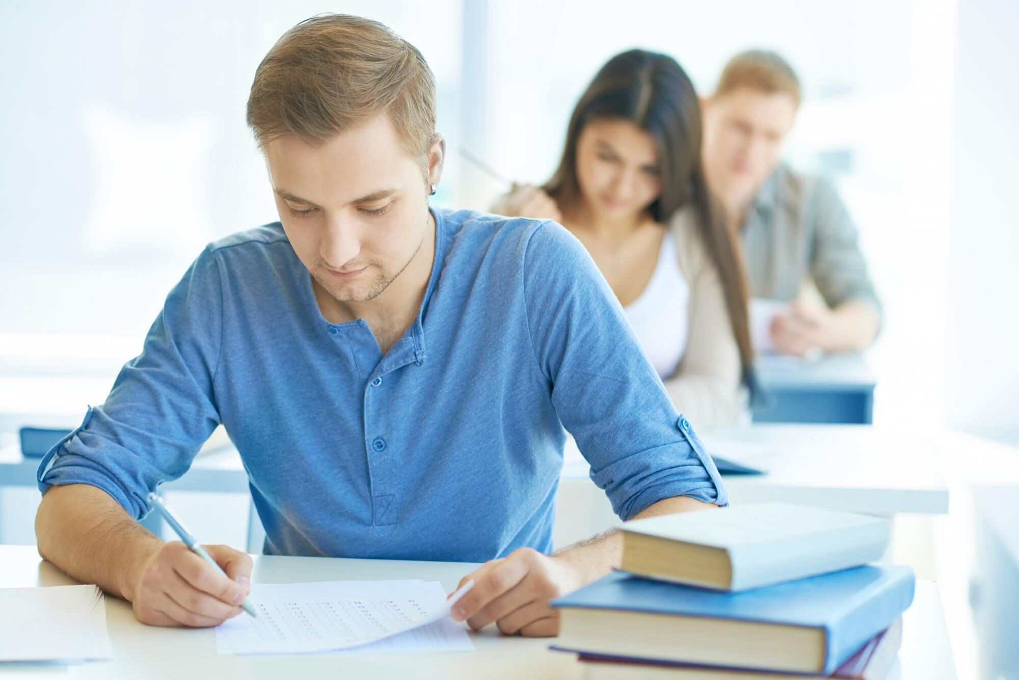 A man writes notes at a desk in a classroom filled with engaged students around him.