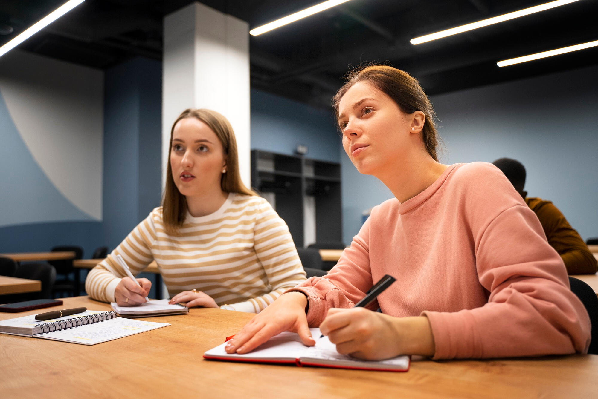 Two young women engaged in conversation while sitting at a table in a classroom setting.
