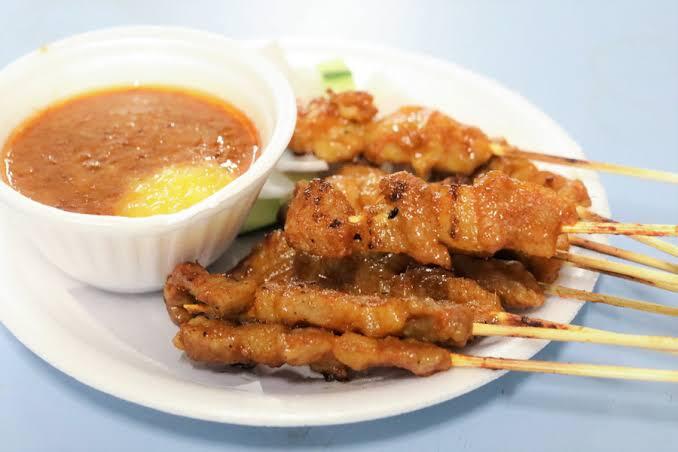 Grilled meat satay skewers on a white plate with cucumber slices, compressed rice cakes, and two small bowls of spicy peanut dipping sauce with a dollop of pineapple puree.