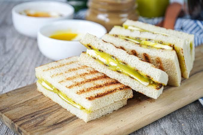 Golden grilled sandwiches with melted cheese and yellow filling are stacked on a wooden board, with dipping bowls and a honey jar behind.