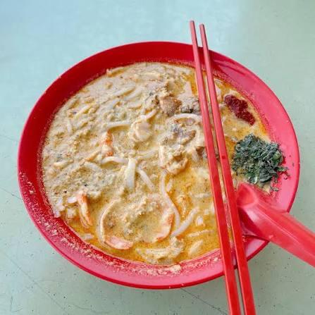 A red bowl of katong laksa with creamy broth, red chopsticks, a spoon, and garnishes on the side, set on a light-colored table.