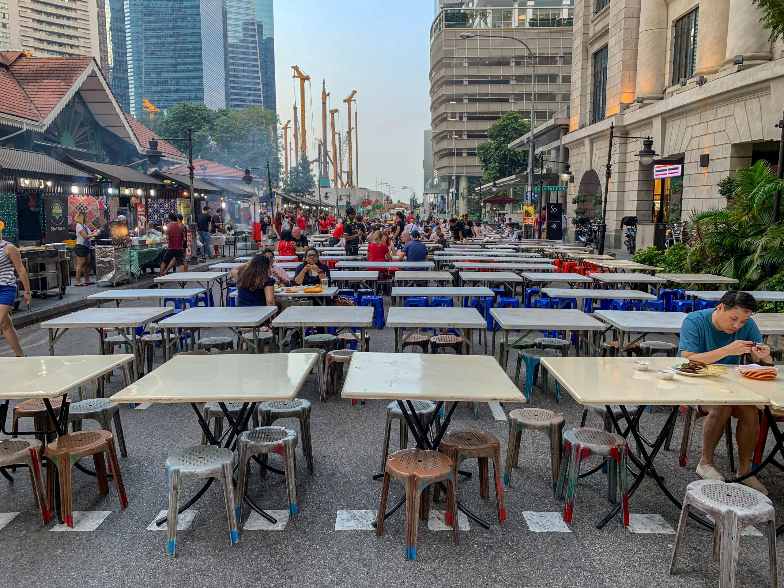 An outdoor street dining area with many white tables and plastic stools, surrounded by city buildings and construction cranes in the background.
