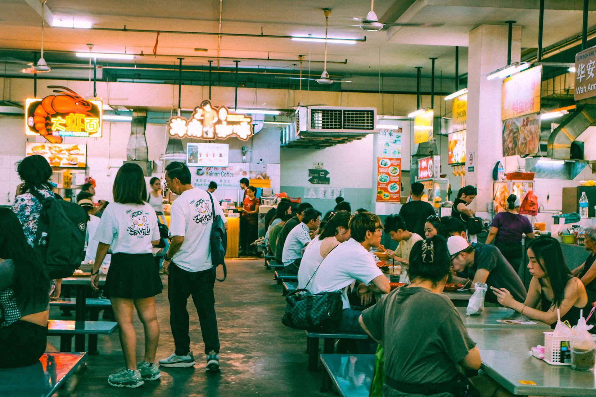 People dine at tables in a busy indoor food court, featuring various food stalls with colorful neon signs and hanging lights under a high ceiling.