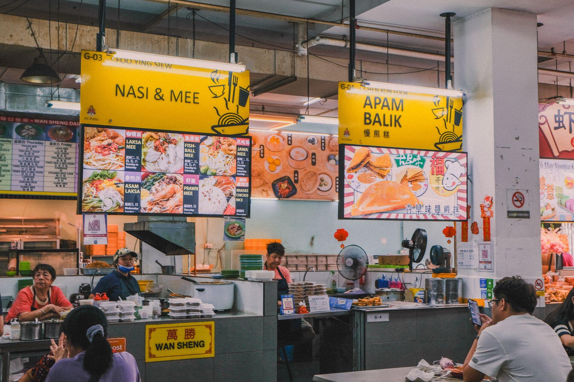 Lively indoor scene of an Asian food court with various stalls and local patrons enjoying meals.