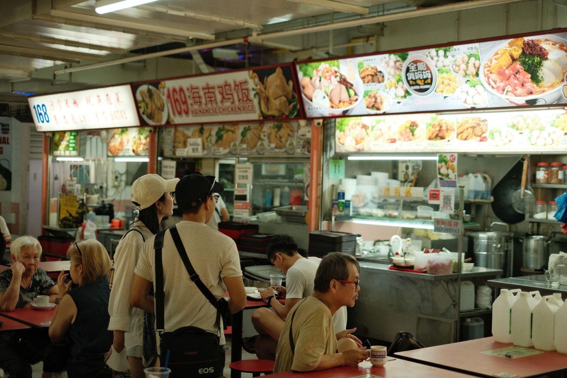 People dine at a food court with stalls displaying menus featuring Hainanese chicken rice and various dishes under bright overhead fluorescent lights.