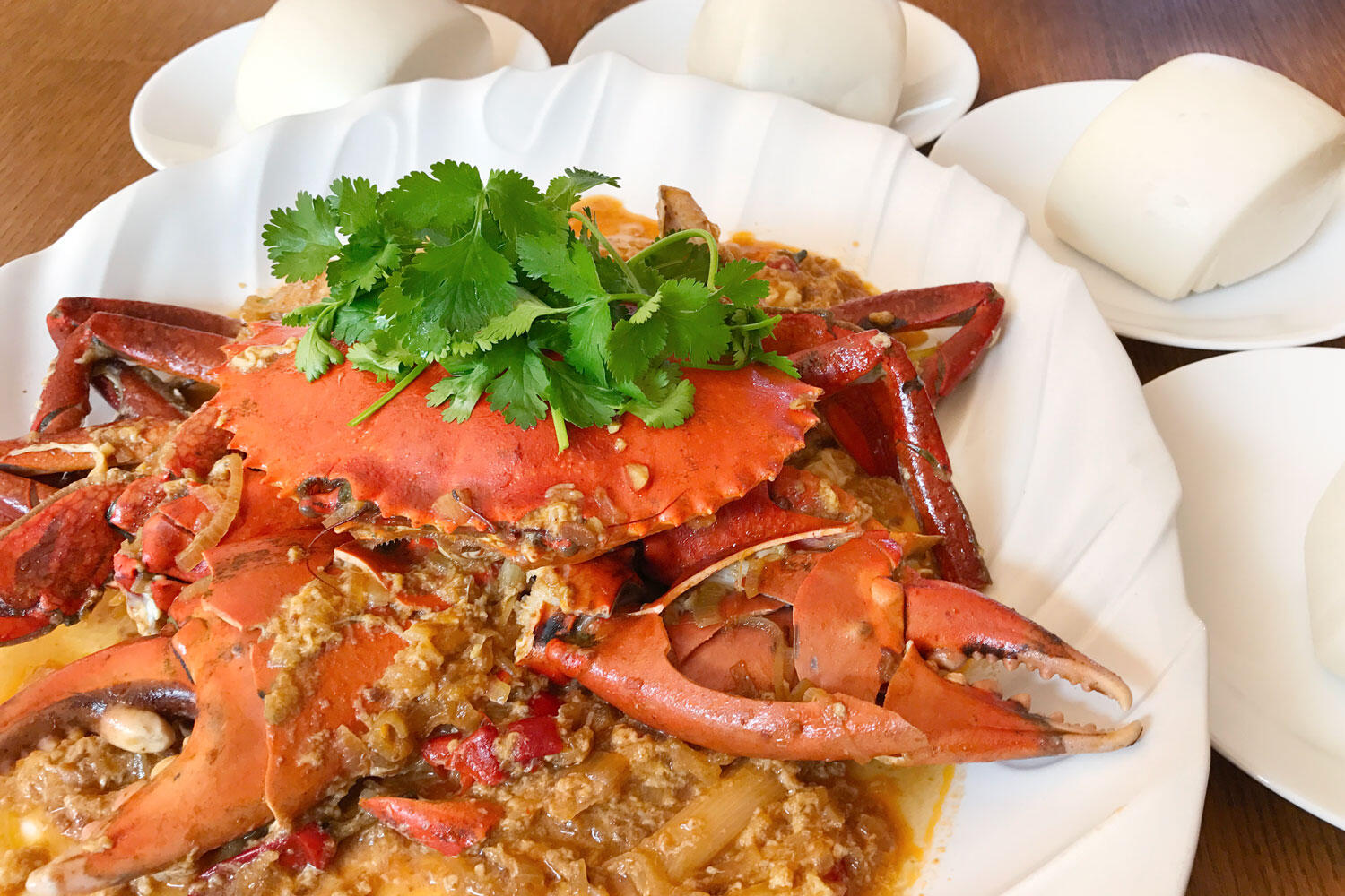 Close-up of spicy chili crab in savory sauce, topped with fresh cilantro, served on a white platter with steamed buns.