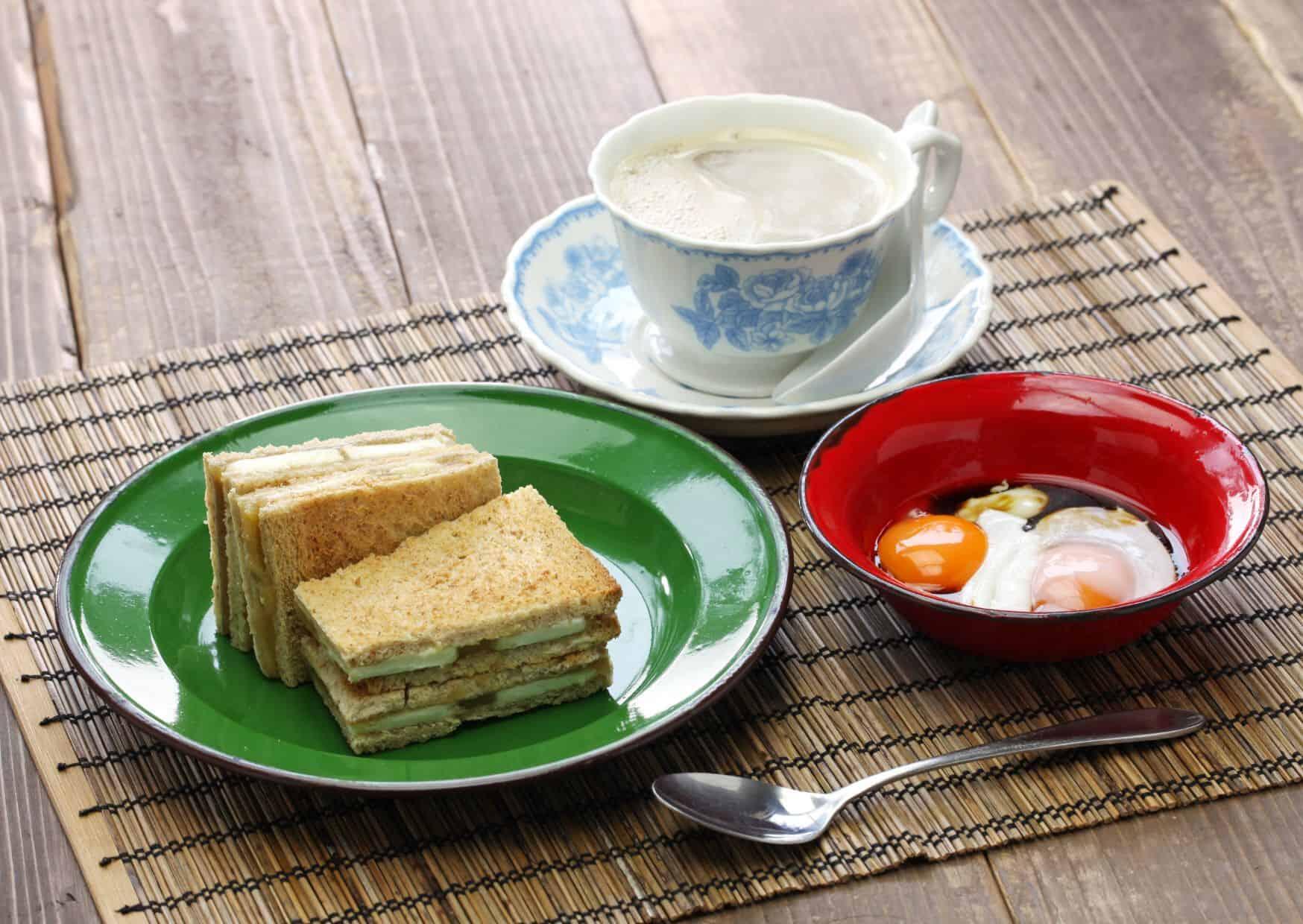 Breakfast setting with stacked sandwiches on a green plate, fried eggs in a red bowl, and coffee in a floral cup on a rustic table.
