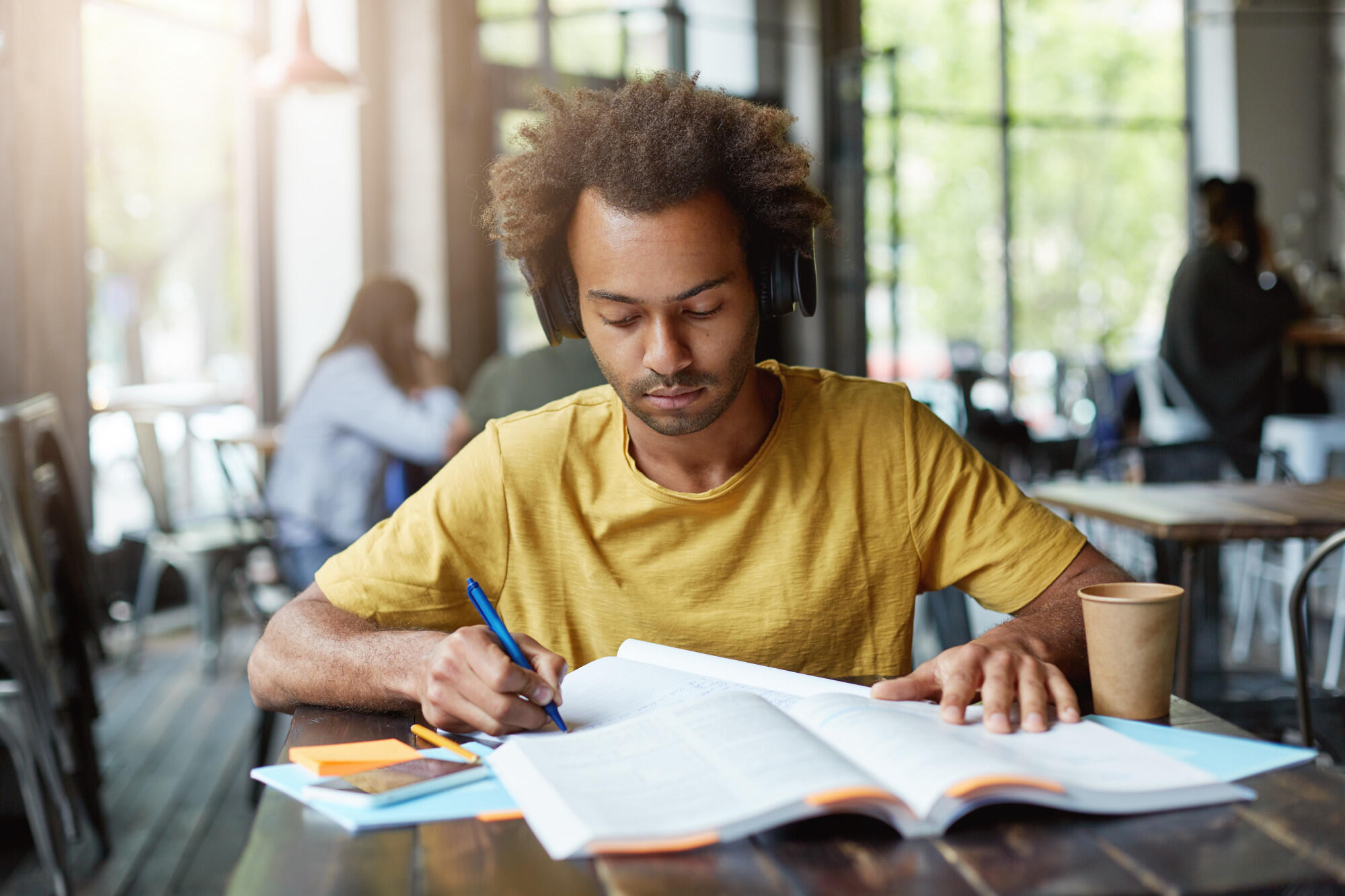 A man wearing headphones studies intently at a table, surrounded by books and notes.