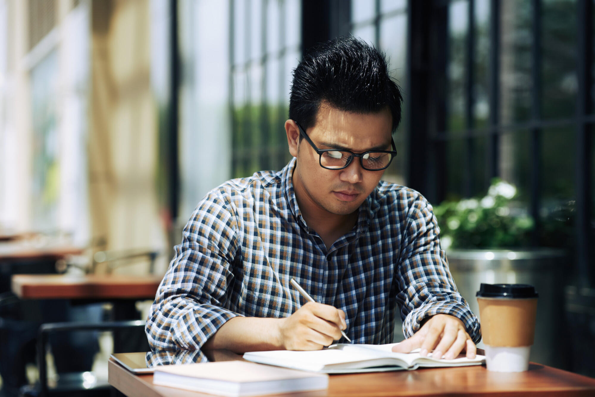 A bespectacled man diligently writes notes in a book, surrounded by a calm environment.