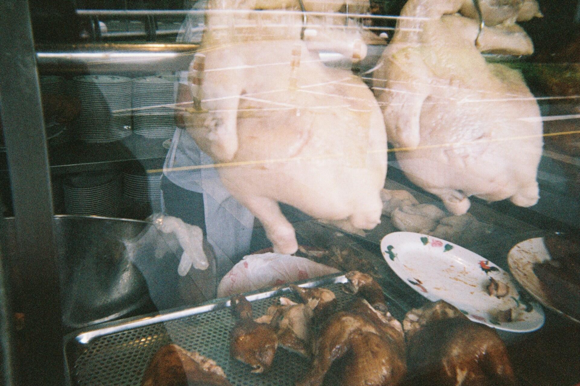 Two whole chickens are hanging above a tray of cooked meat and a plate with food scraps, behind a glass display case.