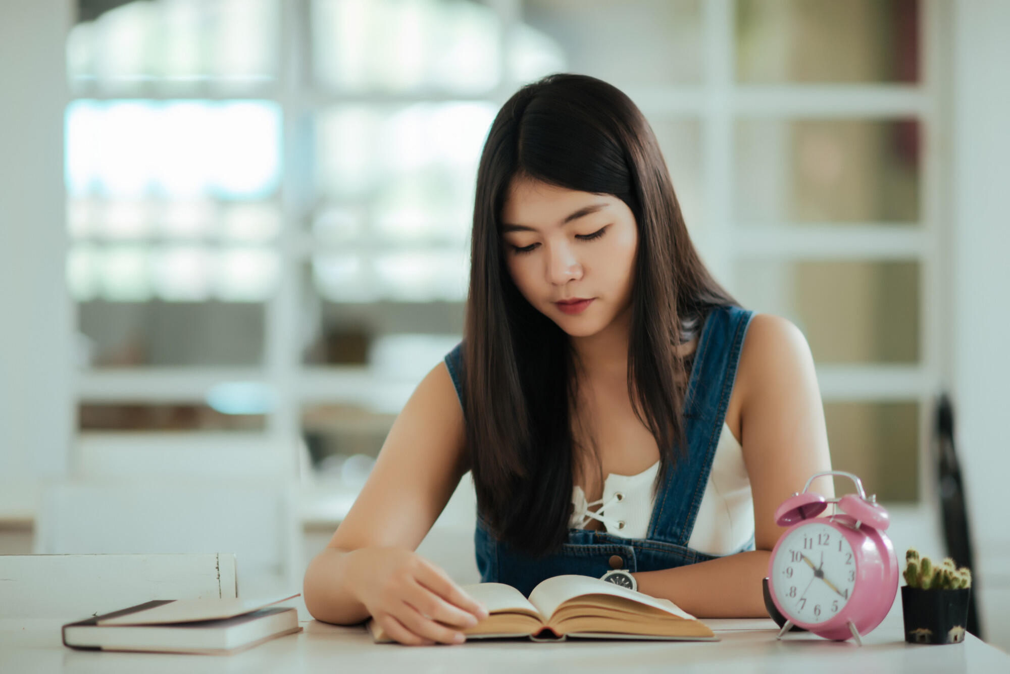 A woman sits at a table, reading an open book, with an alarm clock placed beside her.