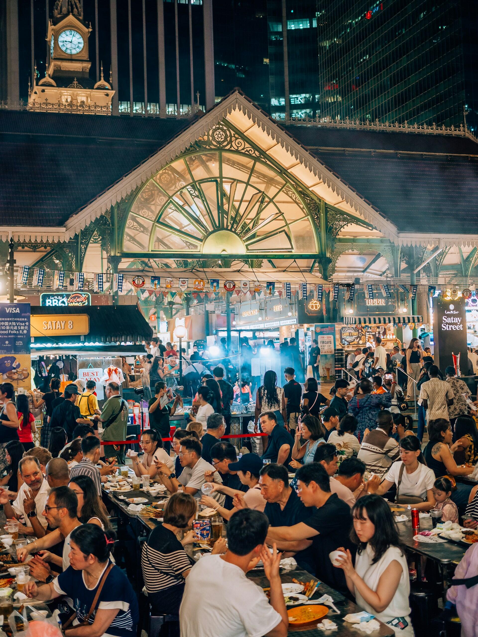 Crowded outdoor night market at Lau Pa Sat in Singapore, featuring people dining at tables under a historic Victorian-style ornate metal roof structure.