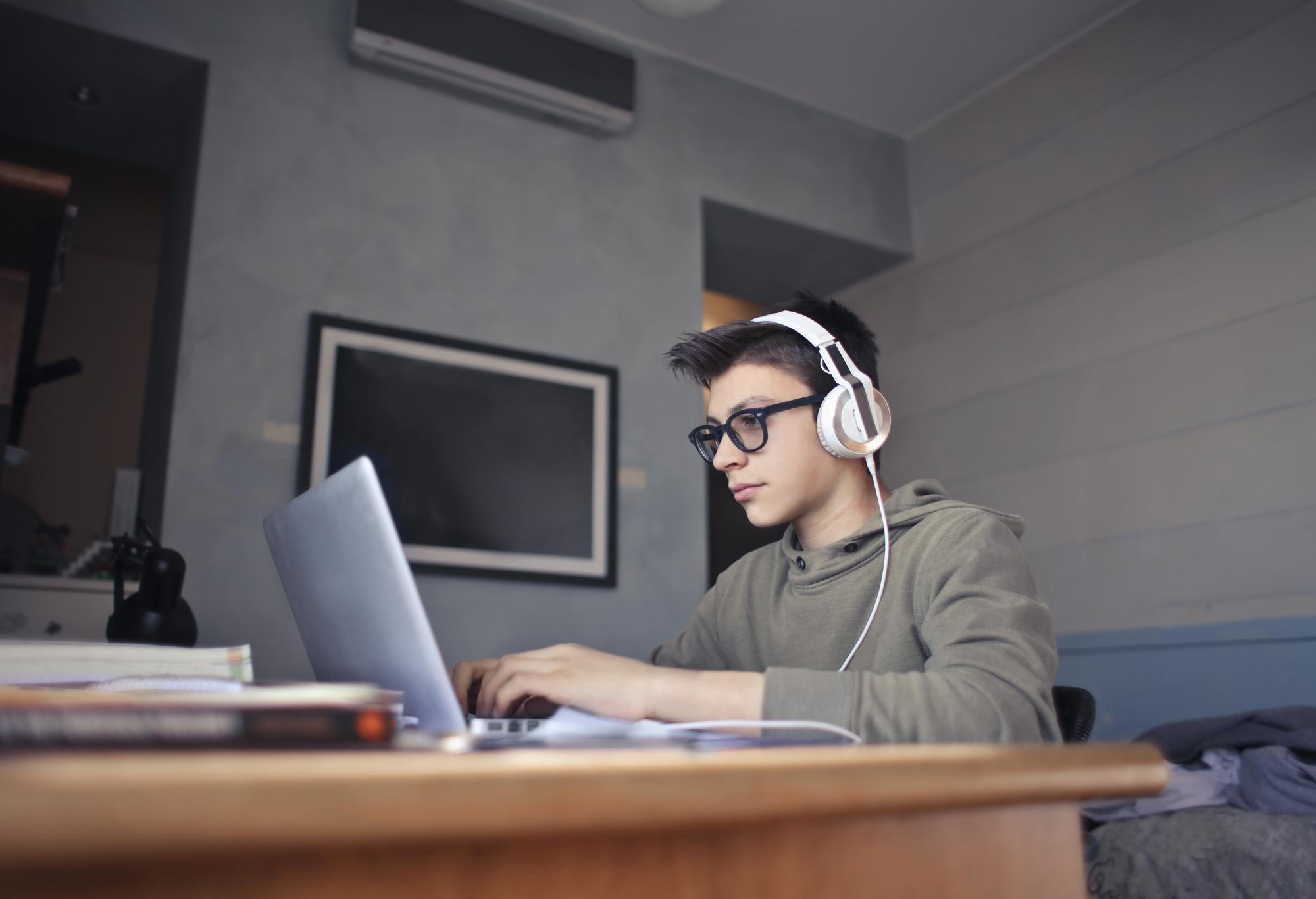Teenage boy sitting at a desk, focused on using a laptop in a cozy home environment.