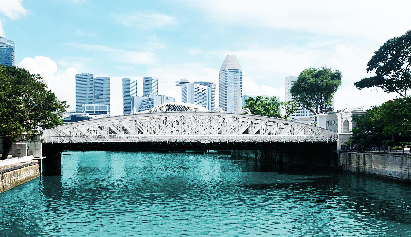 Wide view of the historic white metal arch Anderson Bridge spanning across the Singapore River, with modern city skyscrapers in the background.