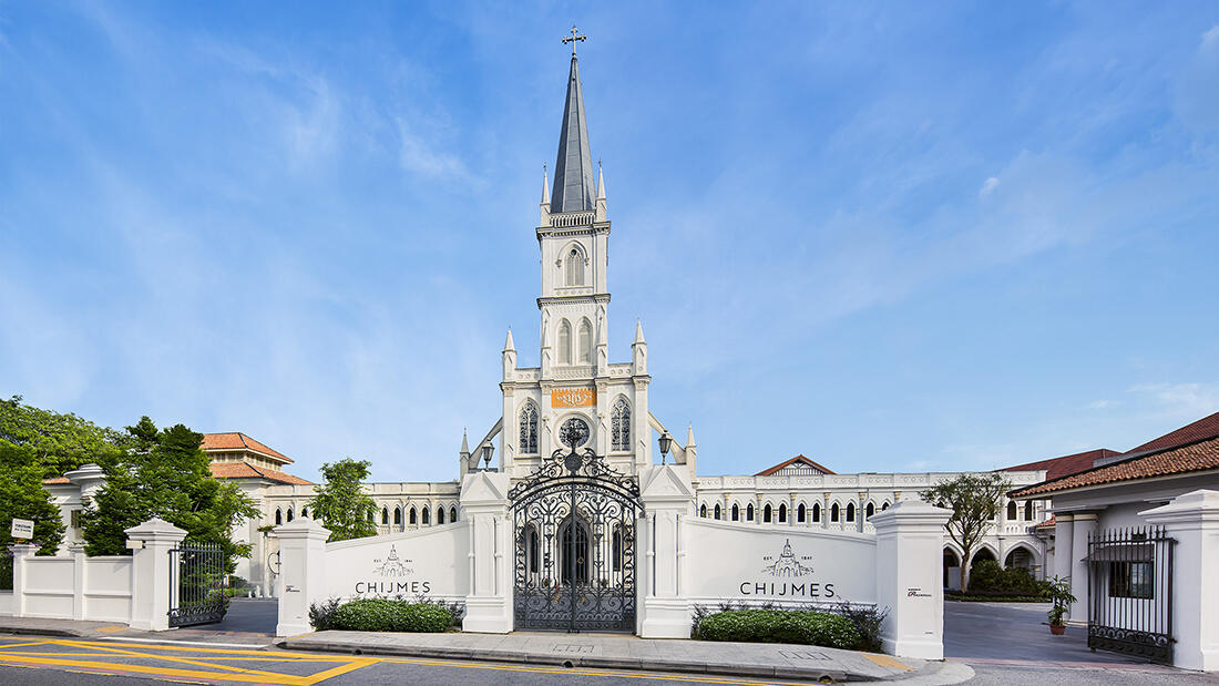 The historic white Gothic-style CHIJMES chapel in Singapore features a tall central spire, arched windows, and an ornate black iron entrance gate.