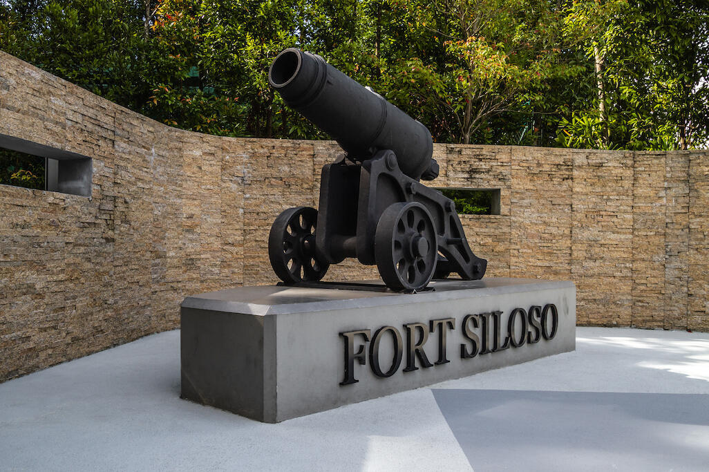 A black vintage cannon sits on a concrete pedestal labeled Fort Siloso, set against a curved stone wall and lush green trees in the background.
