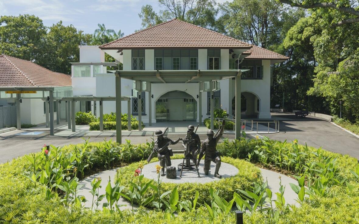 A peaceful garden memorial features bronze sculptures of three soldiers around a table, with lush greenery and a red-roofed building behind them.