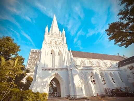 Low-angle exterior view of St. Andrew's Cathedral in Singapore showing its bright white Neo-Gothic facade, tall central spire, and vaulted arches against a bright blue sky.