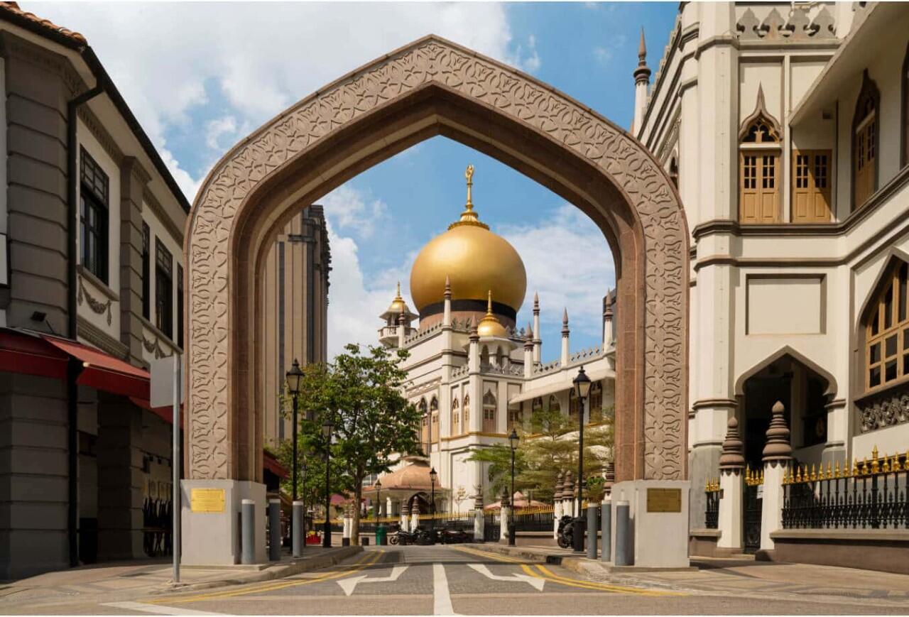 Low-angle view of Sultan Mosque in Singapore featuring its iconic massive golden dome topped with a crescent and star, white exterior walls, minarets, and arched windows.