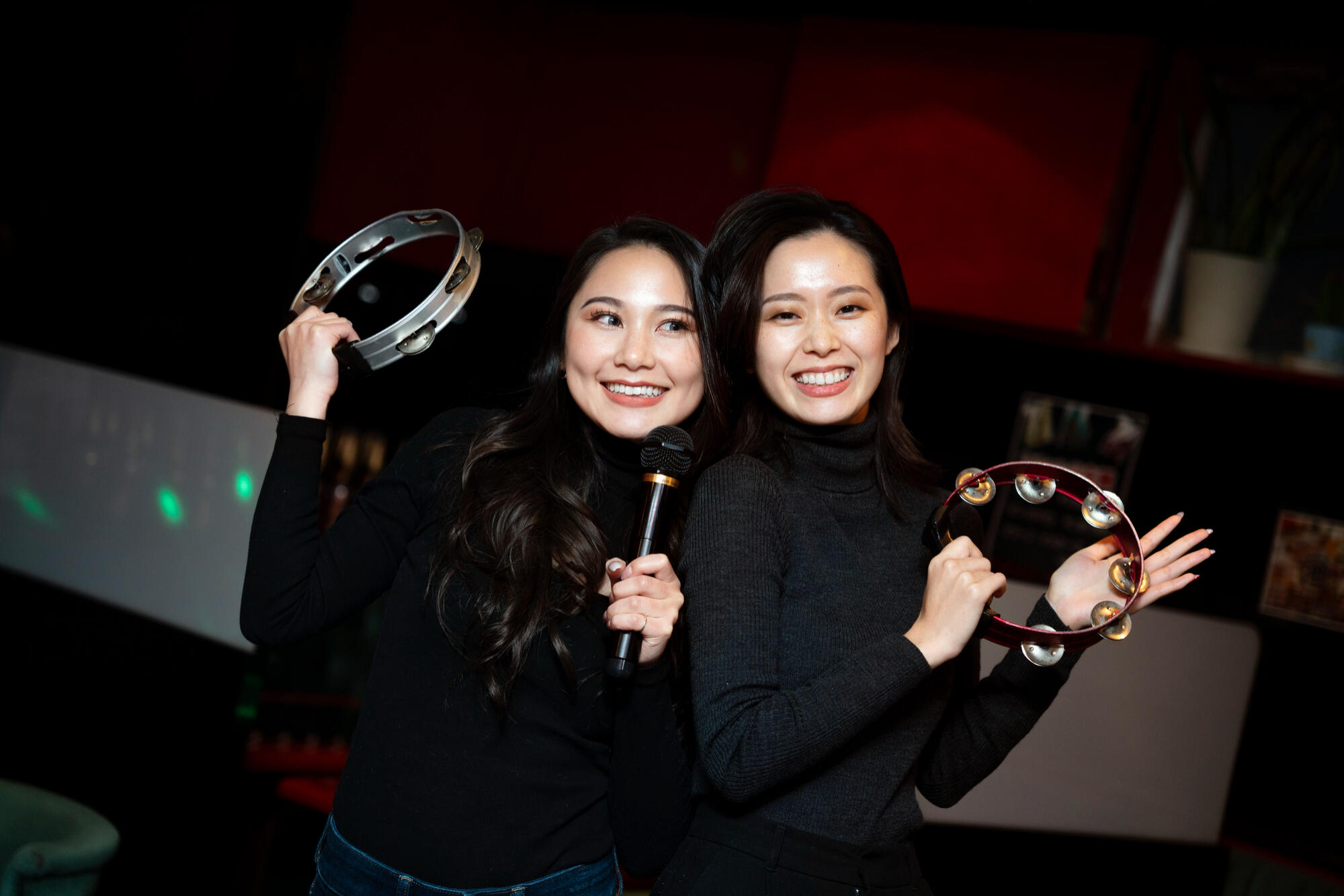 Two Asian women smiling while holding up a pair of tambourines in a lively setting.